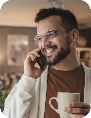 Vīrietis ar brillēm brūnā kreklā un baltā jakā runā pa mobilo telefonu, rokā turot baltu krūzi.