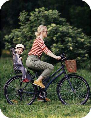 Sieviete sarkanā svītrainā kreklā un zaļās biksēs brauc ar melnu velosipēdu pa zālienu, aizmugurē velosipēda krēsliņā ved mazu bērnu, kuram galvā ir ķivere.
