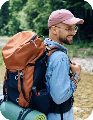 Vīrietis brillēs, rozā keponā un zilā džinsu kreklā, ar oranžu ceļojuma mugursomu un zaļu paklājiņu uz muguras, stāv pie meža malas.