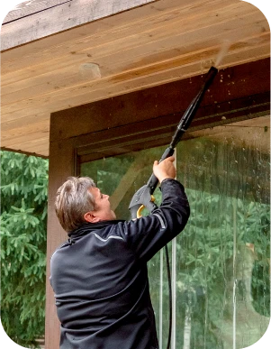 Cilvēks tumšās drēbes ar augstspiediena mazgātāju tīra koka jumtu.