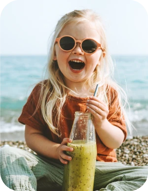 Maza meitene ar gariem gaišiem matiem un saulesbrillēm uz acīm sēž pludmalē un dzer zaļu dzērienu no stikla pudeles caur salmiņu.