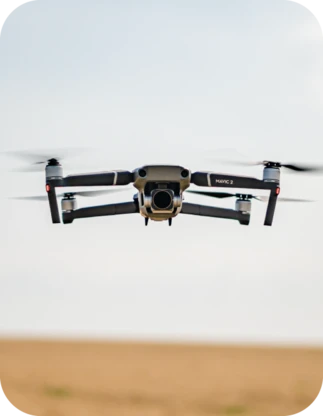 Pelēks Mavic 2 drons ar kameru lido gaisā virs lauka.