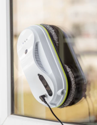 Automātisks logu tīrīšanas robots baltā krāsā piestiprināts pie stikla loga no iekšpuses, tuvplānā ovāla ierīce ar ventilācijas atverēm, indikatoru gaismām un pieslēgtu barošanas kabeli.
