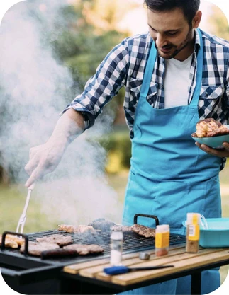 Vīrietis zilā priekšautā un rūtainā kreklā grilē gaļu uz āra grila, turot šķīvi ar gaļas gabaliem vienā rokā un knaibles otrā, fonā zaļā zāliena teritorija.