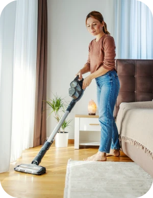 Sieviete tīra koka grīdu pie gultas ar rokas bezvadu vertikālo putekļsūcēju. Fonā gaišs paklājs, balti aizkari, telpaugs, gaišs nakts skapītis un liela gulta.