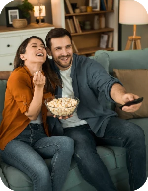 Sieviete un vīrietis sēž mājīgā viesistabā uz dīvāna, ēd popkornu no stikla bļodas un skatās televizoru. Sieviete tur popkorna bļotu, bet vīrietis izstieptā rokā tur televizora pulti.