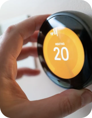 Cilvēka roka regulē melnu apaļu termostatu ar oranžu ekrānu, kurā rakstīts 'HEATING' un temperatūra 20 °C.