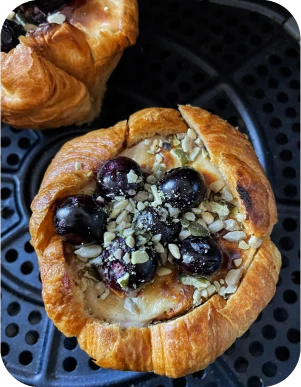 Kruasāna groziņš pildīts ar krēmīgu masu, virsū svaigas tumšas ogas un sēkliņu maisījums, novietots uz gaisa fritera restes.