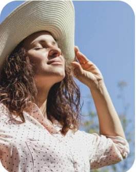 Sieviete ar cirtainiem matiem gaišā cepurē un punktainā krekliņā stāv ārā zem zilām debesīm, viņas seju apspīd saules stari.