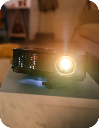 Mājas vidē uz galda novietots ieslēgts projektors ar spilgtu gaismas staru, apkārt silti apgaismotam interjeram.