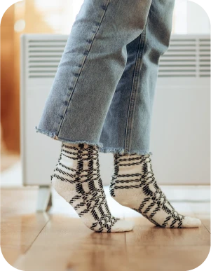 Cilvēks baltās zeķēs ar melnu svītru rakstu un zilās džinsu biksēs stāv uz koka grīdas pie balta elektriskā radiatora.