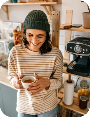 Sieviete zaļā adītā cepurē un svītrainā džemperī tur baltu krūzi, stāvot virtuvē pie kafijas automāta.