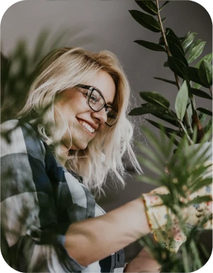 Sieviete ar gaišiem matiem, brillēm, rūtainā kreklā un dzeltenos cimdos ar ziedu rakstu kopj zaļus istabas augus, fonā telpa ar daudz augiem.