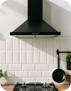 Virtuve ar melnu tvaika nosūcēju, baltiem flīzētiem sienas paneļiem, gāzes plīti un pie sienas piekarinātu pannu.