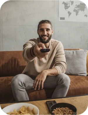 Vīrietis gaišās drēbēs sēž uz dīvāna ar televizora pulti rokā; priekšplānā galds ar čipsiem un riekstiem, fonā pie sienas pasaules karte.