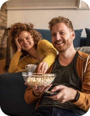 Sieviete un vīrietis sēž uz dīvāna un skatās televizoru; vīrietis tur bļodu ar popkornu, sieviete apgūlusies dīvāna atspiež galvu uz vienas rokas, bet ar otru sniedzas pēc popkorna.