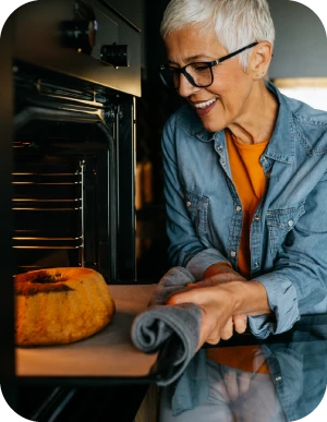 Sieviete ar brillēm, zilā džinsa kreklā, stāv pie atvērtas cepeškrāsns un ņem ārā svaigi ceptu kēksu, turot cepeškrāsns pannu ar dvieli.