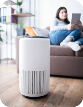 Balts cilindrisks bezvadu skaļrunis uz brūnas koka grīdas, fonā sieviete ar planšetdatoru rokās sēž uz tumši brūna dīvāna.