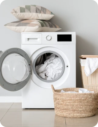 Balta veļas mašīna ar atvērtām durvīm un baltu veļu tajā, blakus divi veļas grozi ar gaišu veļu; uz veļas mašīnas virsmas novietoti divi dekoratīvi spilveni.