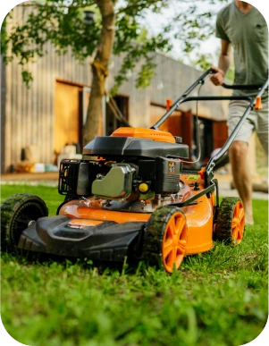 Vīrietis gaišās drēbēs pļauj zālienu ar oranžas krāsas zāles pļāvēju dārzā, fonā pelēka koka ēka un koki.