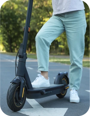 Cilvēks gaiši zilās biksēs un baltās kedās stāv vienu kāju uzlicis uz elektriskā skrejriteņa un otru blakus tam, uz asfaltēta ceļa ar baltu, lielu bultu, fonā koki.