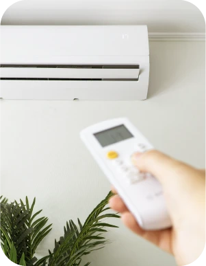 Persona ar baltu tālvadības pulti vērstu pret sienas gaisa kondicionieri, fonā zaļš istabas augs.