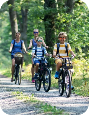 Četri cilvēki brauc ar velosipēdiem pa meža taku — priekšplānā divi bērni svītrainos kreklos, fonā divi pieaugušie, apkārt zaļa mežaina vide.