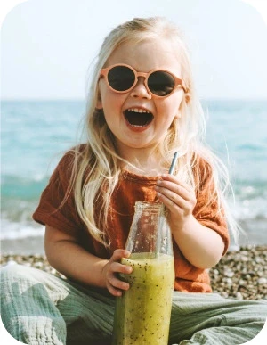 Maza meitene ar gariem gaišiem matiem un saulesbrillēm uz acīm sēž pludmalē un dzer zaļu dzērienu no stikla pudeles caur salmiņu.