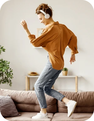 Sieviete dzeltenā kreklā, džinsos, baltās kedās un ar baltām austiņām ap galvu dejo uz brūna dīvāna dzīvojamā istabā.