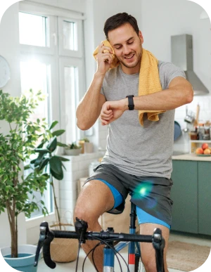 Vīrietis pelēkā T-kreklā un šortos uz velo trenažiera ar dzeltenu dvieli ap pleciem un skatās uz viedpulkstenī; fonā virtuve ar augiem un logu.