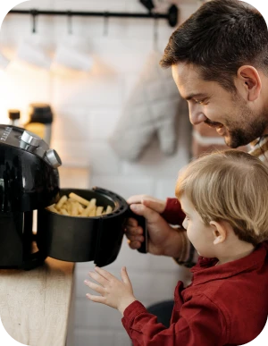 Vīrietis un bērns virtuvē kopā gatavo — pieaugušais tur rokās aerogrilu pilnu ar frī kartupeļiem, bērns ar interesi skatās.