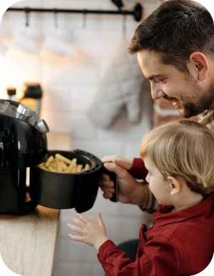 Vīrietis un bērns virtuvē kopā gatavo — pieaugušais tur rokās aerogrilu pilnu ar frī kartupeļiem, bērns ar interesi skatās.