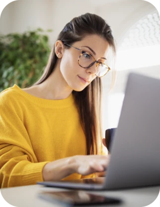 Sieviete ar gariem matiem, brillēm un dzeltenā džemperī strādā pie klēpjdatora, vienā rokā turot kafijas krūzi.