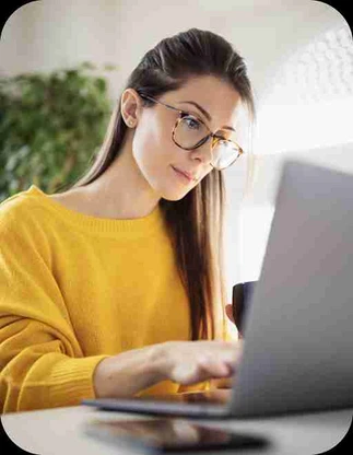 Sieviete ar gariem matiem, brillēm un dzeltenā džemperī strādā pie klēpjdatora, vienā rokā turot kafijas krūzi.