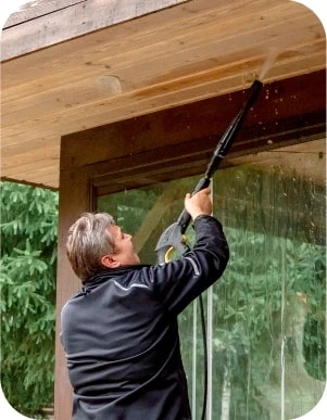 Cilvēks tumšās drēbes ar augstspiediena mazgātāju tīra koka jumtu.