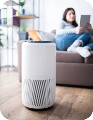 Balts cilindrisks bezvadu skaļrunis uz brūnas koka grīdas, fonā sieviete ar planšetdatoru rokās sēž uz tumši brūna dīvāna.