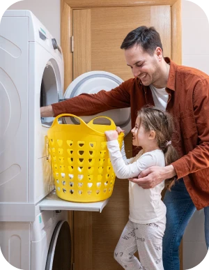 Tēvs un meita kopīgi izņem veļu no priekšējās ielādes veļas žāvētājā un ievieto dzeltenā veļas grozā. Veļas žāvētājs ir novietots virs veļas mašīnas.