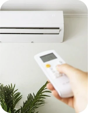 Persona ar baltu tālvadības pulti vērstu pret sienas gaisa kondicionieri, fonā zaļš istabas augs.
