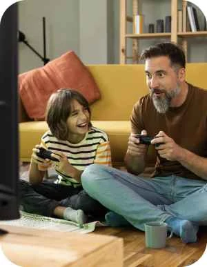 Vīrietis un bērns sēž uz grīdas dzīvojamā istabā un spēlē videospēles ar kontrolieriem, fonā dzeltens dīvāns.