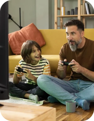 Vīrietis un bērns sēž uz grīdas dzīvojamā istabā un spēlē videospēles ar kontrolieriem, fonā dzeltens dīvāns.