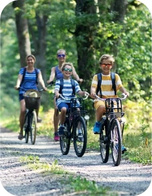 Četri cilvēki brauc ar velosipēdiem pa meža taku — priekšplānā divi bērni svītrainos kreklos, fonā divi pieaugušie, apkārt zaļa mežaina vide.