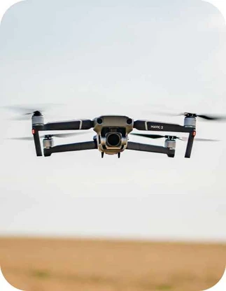 Pelēks Mavic 2 drons ar kameru lido gaisā virs lauka.