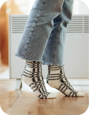 Cilvēks baltās zeķēs ar melnu svītru rakstu un zilās džinsu biksēs stāv uz koka grīdas pie balta elektriskā radiatora.