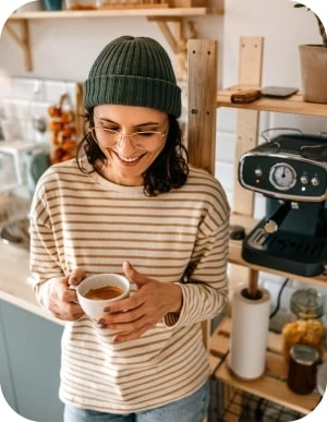 Sieviete zaļā adītā cepurē un svītrainā džemperī tur baltu krūzi, stāvot virtuvē pie kafijas automāta.