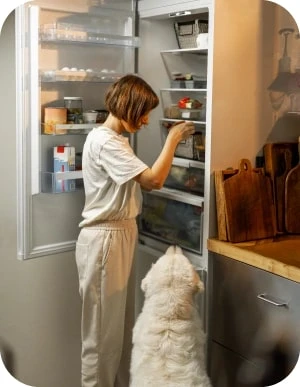 Cilvēks gaišā apģērbā stāv pie atvērta ledusskapja un skatās tajā iekšā, blakus stāv liels, balts suns un skatās uz cilvēku.