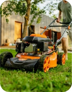 Vīrietis gaišās drēbēs pļauj zālienu ar oranžas krāsas zāles pļāvēju dārzā, fonā pelēka koka ēka un koki.