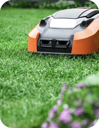 Oranžs zāles pļāvējs robots uz zaļa zāliena ar purpursarkaniem ziediem priekšplānā.