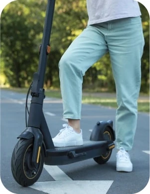 Cilvēks gaiši zilās biksēs un baltās kedās stāv vienu kāju uzlicis uz elektriskā skrejriteņa un otru blakus tam, uz asfaltēta ceļa ar baltu, lielu bultu, fonā koki.