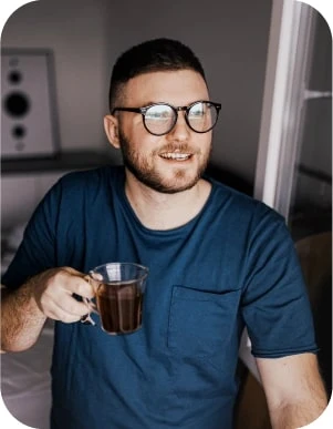 Vīrietis ar brillēm, zilā kreklā tur caurspīdīgu krūzi ar dzērienu, fonā skaļrunis un logs.