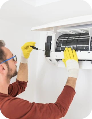 Vīrietis ar brillēm, brūnā apģērbā un dzeltenos cimdos labo gaisa kondicionieri ar skrūvgriezi, kas piestiprināts pie sienas.